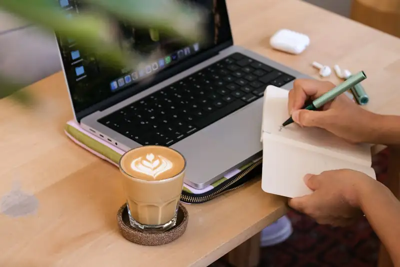 Person writing content plans in a notebook beside a laptop and coffee